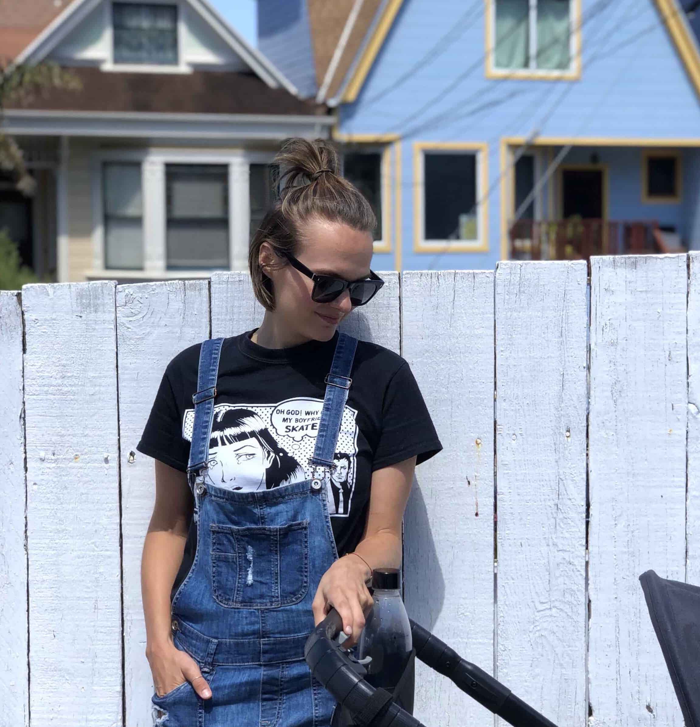Woman in blue jean overalls leaning against a wooden fence with one hand on a stroller looking down
