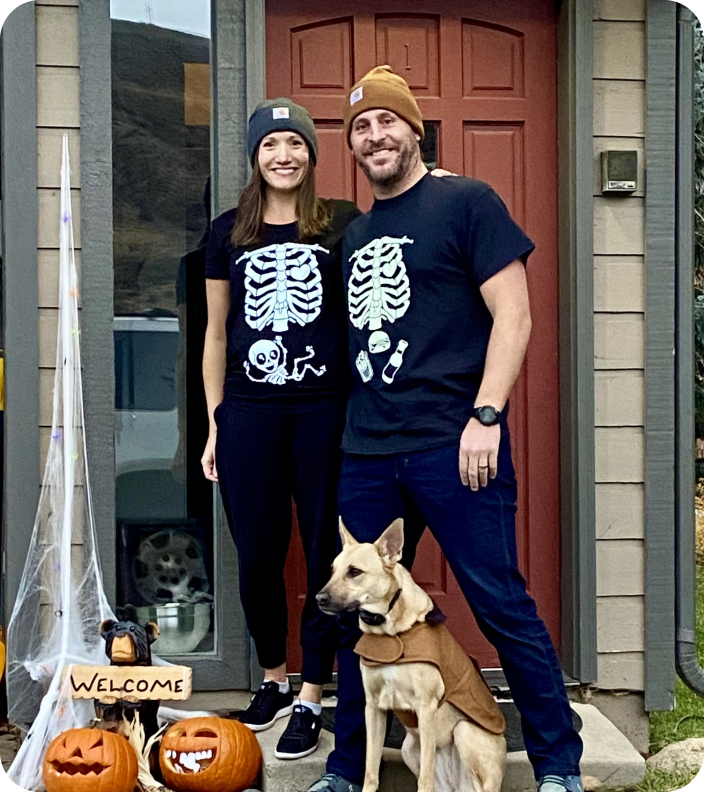 Couple and their dog posing in front of their porch, dressed for Halloween, with a 'welcome' sign and pumpkins