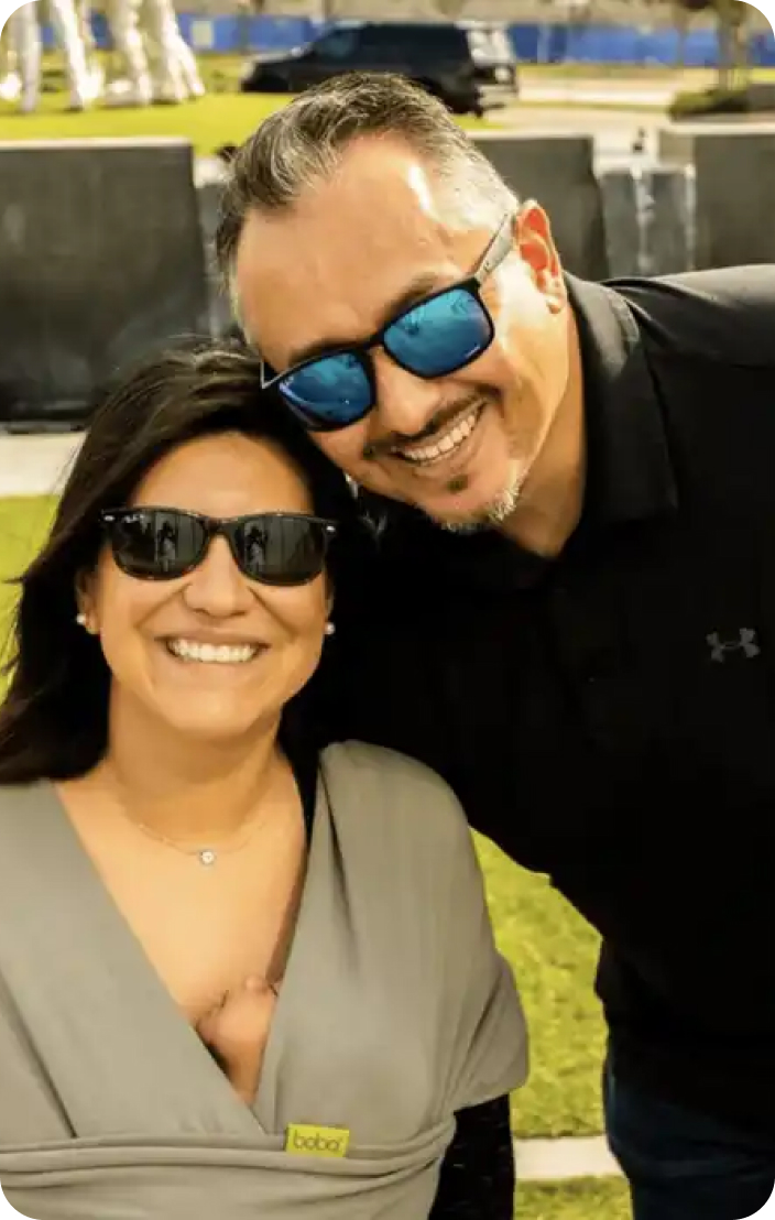 Man and woman in a garden wearing sunglasses, woman carrying her baby in babywearing gear, both smiling