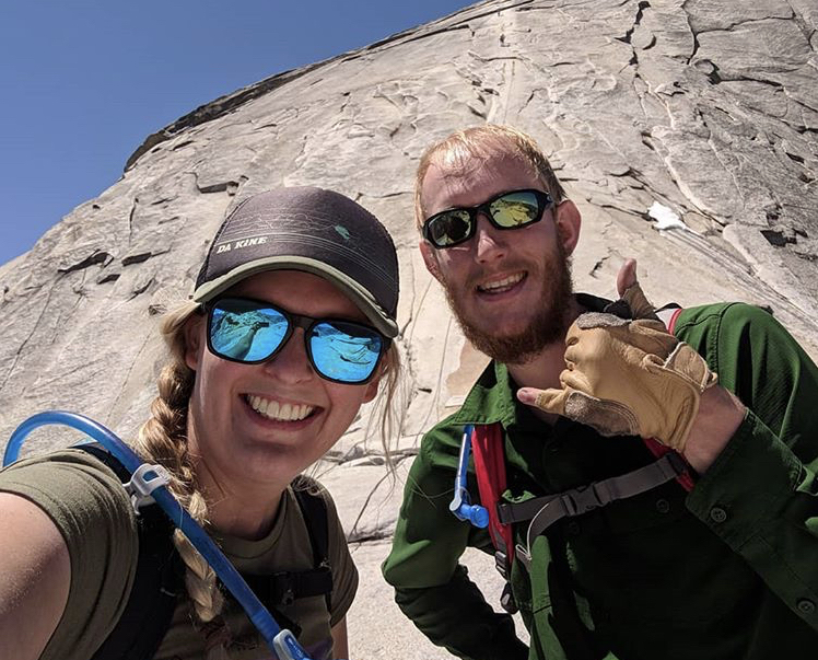 Woman and a man smiling wearing rock climbing gear standing on the side of a mountain